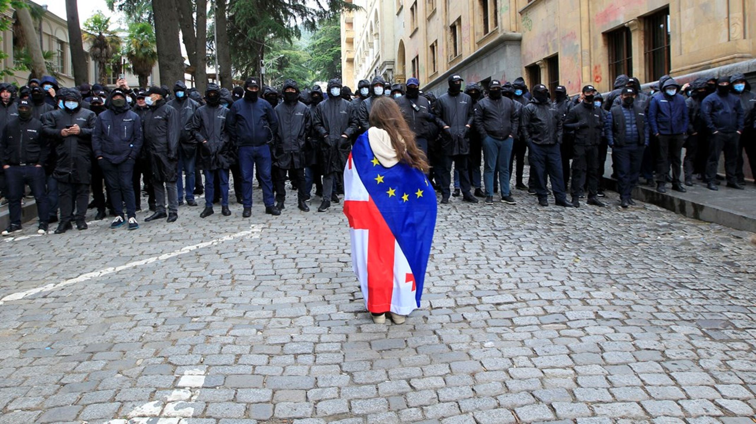 En ung kvinde svøbt i Georgiens og EU's flag står foran maskerede politifolk i hovedstaden Tbilisi tirsdag. Tusinder af borgere har demonstreret mod vedtagelsen af en ny lov, de opfatter som antidemokratisk.